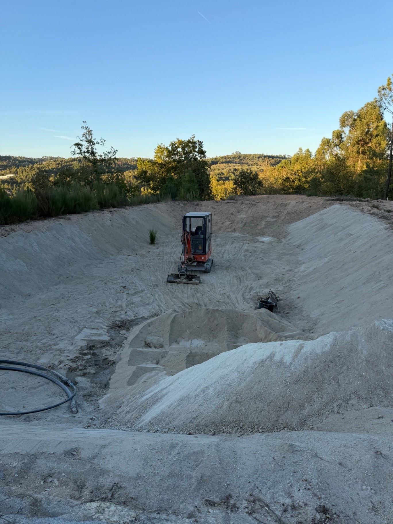 Charca em construção com escavadora no fundo e materiais minerais em primeiro plano